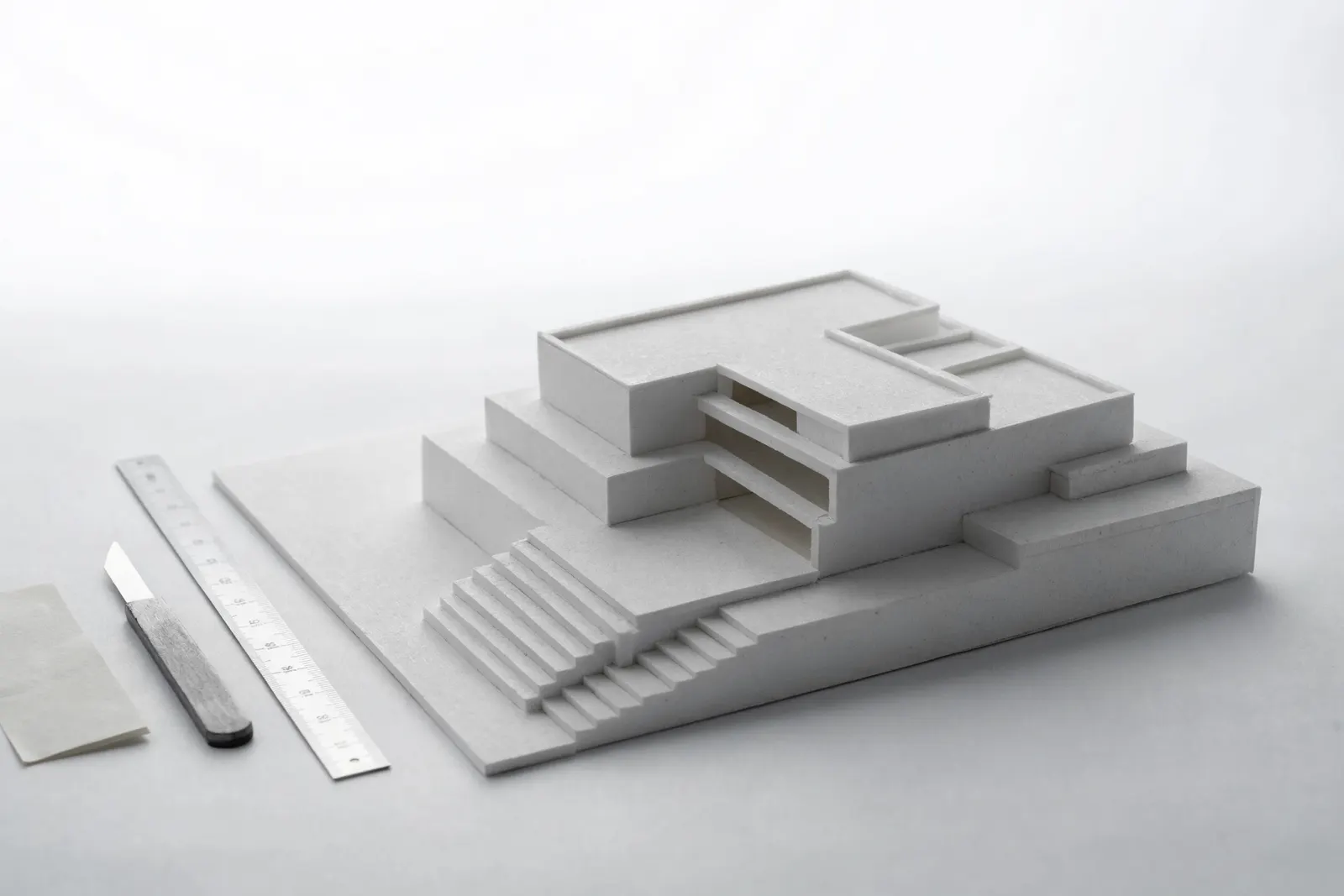 A white architectural model on a white table under even studio light, a scalpel and a coordinate note set beside it.